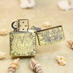 Decorative gold Zippo lighter with intricate engravings, open, resting on sandy beach with seashells nearby.