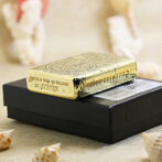 Gold Zippo lighter with ornate engraving resting on a black box, surrounded by seashells on a sandy surface.