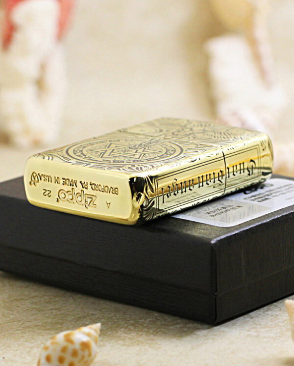 KZ211-05 Gold Zippo lighter with ornate engraving resting on a black box, surrounded by seashells on a sandy surface.