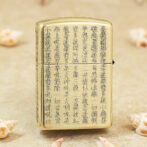Gold Zippo lighter with vertical Chinese script engraved across its front, set on a sandy surface with small seashells nearby, conveying a decorative, cultural motif.