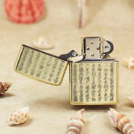Gold Zippo-style lighter with engraved Asian characters, open, resting on sandy beach among seashells.
