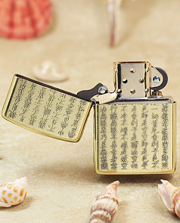 KZ213-02 Gold Zippo-style lighter with engraved Asian characters, open, resting on sandy beach among seashells.
