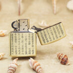 Brass Zippo lighter with engraved Chinese characters, open lid, surrounded by seashells on a sandy surface