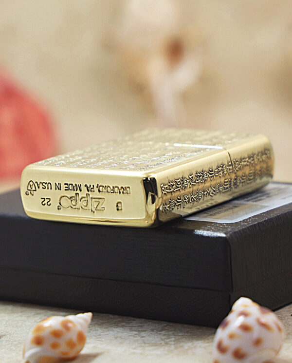 KZ213-05 Gold Zippo lighter with engraved designs resting on a black box, surrounded by seashells and a soft, blurred background.