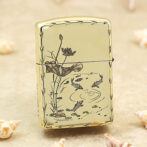 Gold Zippo-style lighter with a koi pond and lotus flower design on a pale background, set on sand with seashells nearby.