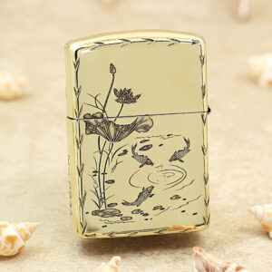 Gold Zippo-style lighter with a koi pond and lotus flower design on a pale background, set on sand with seashells nearby.