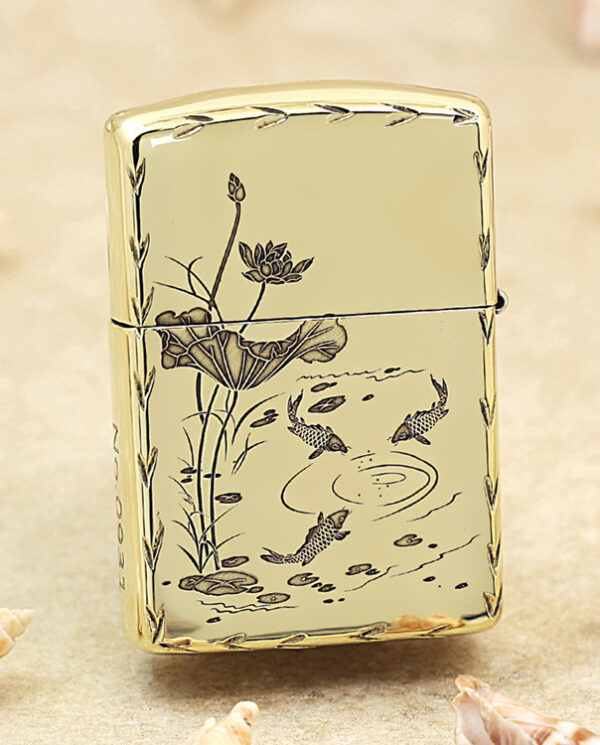 KZ214-01 Gold Zippo-style lighter with a koi pond and lotus flower design on a pale background, set on sand with seashells nearby.