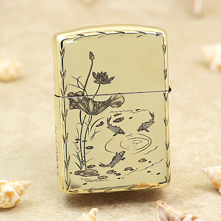 Gold Zippo-style lighter with a koi pond and lotus flower design on a pale background, set on sand with seashells nearby.