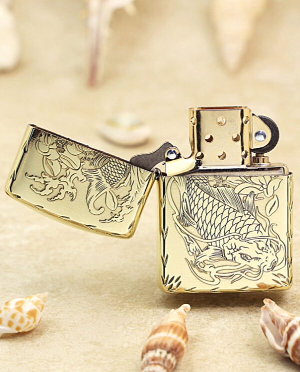 KZ214-02 Gold engraved dragon lighter with open lid on a sandy beach, seashells nearby.