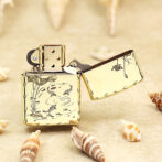 Gold-toned lighter with an open lid, featuring engraved koi fish and plant motifs, surrounded by seashells on a sandy surface.