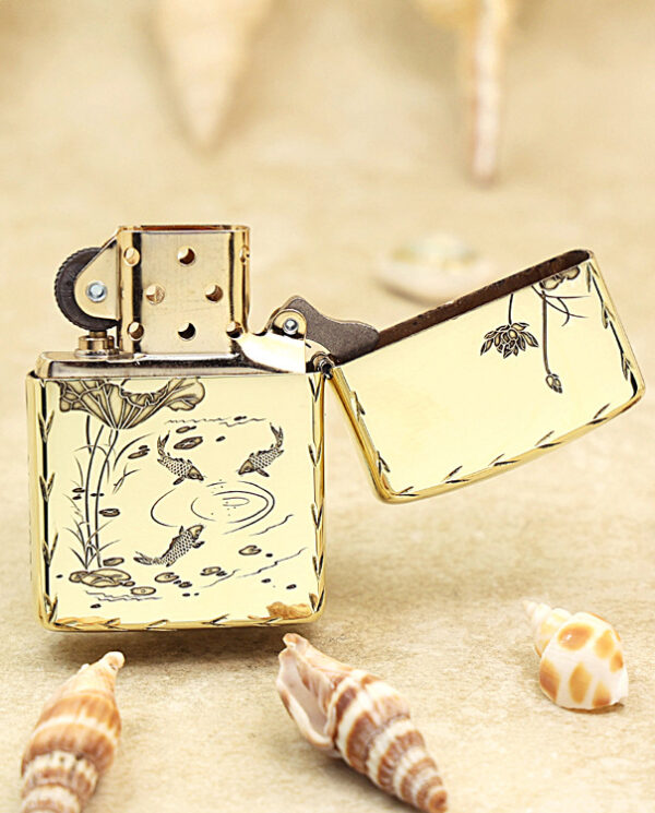 KZ214-03 Gold-toned lighter with an open lid, featuring engraved koi fish and plant motifs, surrounded by seashells on a sandy surface.