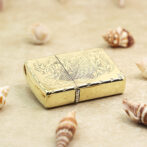 Gold metal lighter with an engraved fish design resting on sand among seashells on a beach-inspired surface.