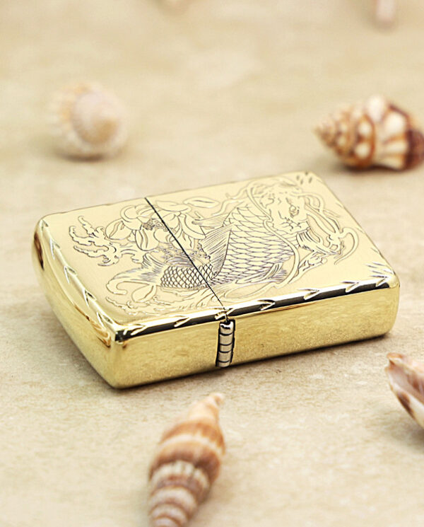 KZ214-04 Gold metal lighter with an engraved fish design resting on sand among seashells on a beach-inspired surface.