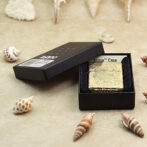 Gold engraved Zippo lighter in a black display box on a sandy surface with assorted seashells nearby.