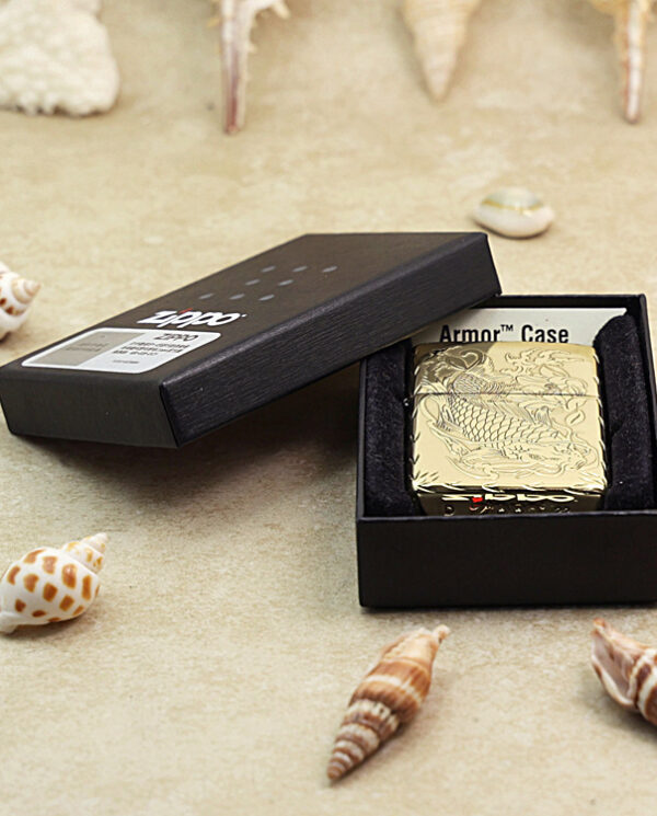 KZ214-06 Gold engraved Zippo lighter in a black display box on a sandy surface with assorted seashells nearby.