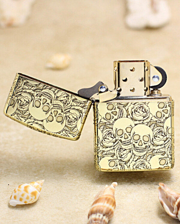 KZ215-02 Gold Zippo-style lighter with skull and rose engraving, open lid, resting on sandy beach with seashells nearby.