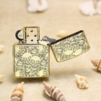 Gold lighter with skull pattern and open lid resting on sand among seashells.