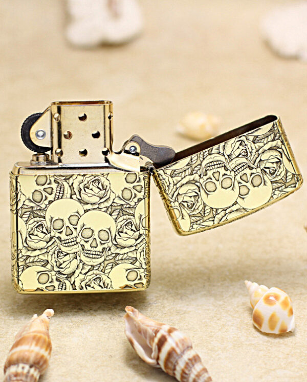 KZ215-03 Gold lighter with skull pattern and open lid resting on sand among seashells.