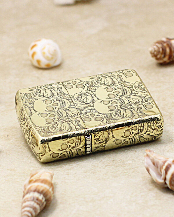 KZ215-04 Gold patterned lighter with skulls, placed on sandy surface among seashells.