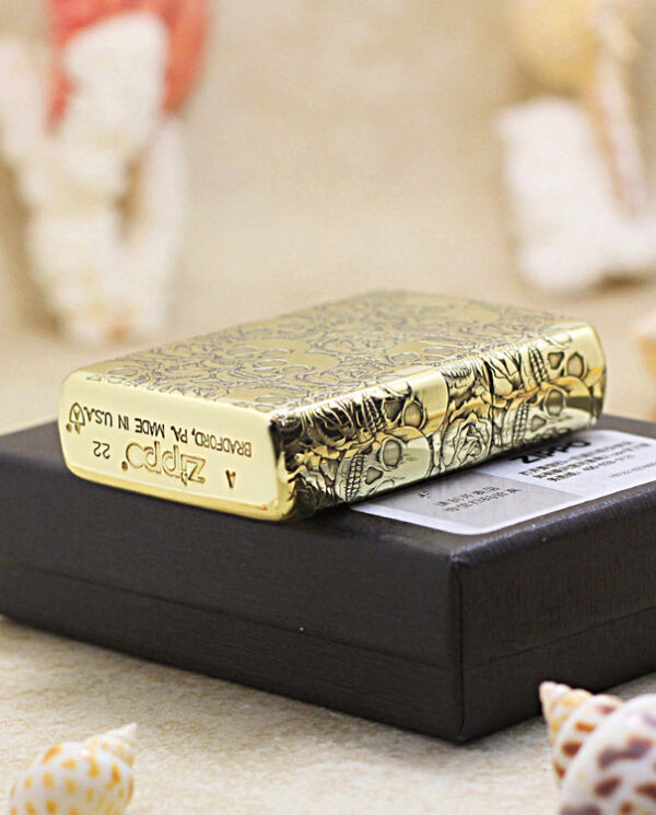 KZ215-05 Ornate gold Zippo-style lighter resting on a black box, with seashells in the background.