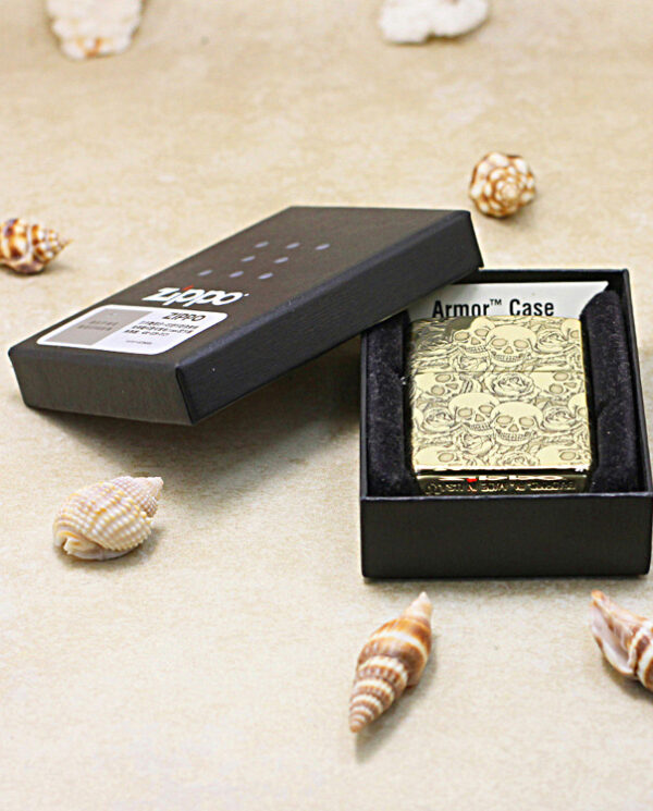 KZ215-06 Gold skull-pattern lighter in a black presentation box, surrounded by seashells on a sandy surface.
