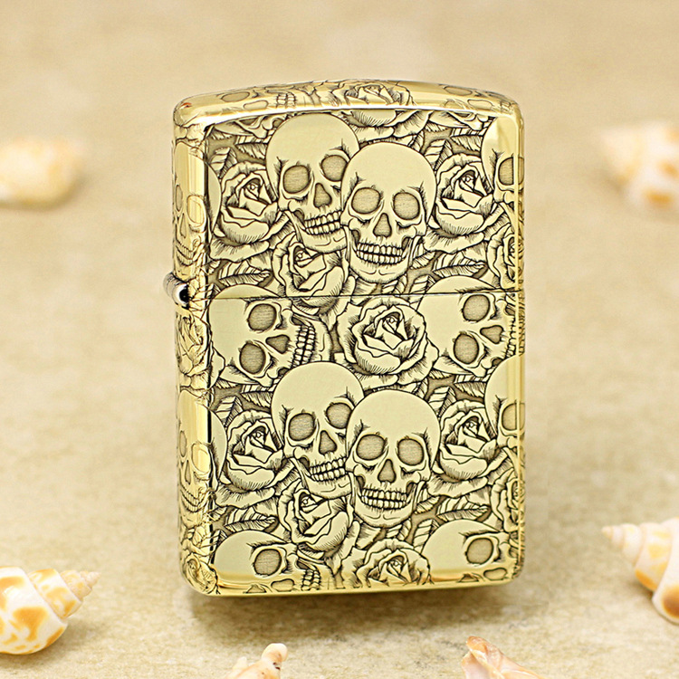 Gold Zippo lighter covered in skull and rose engravings on a sandy surface