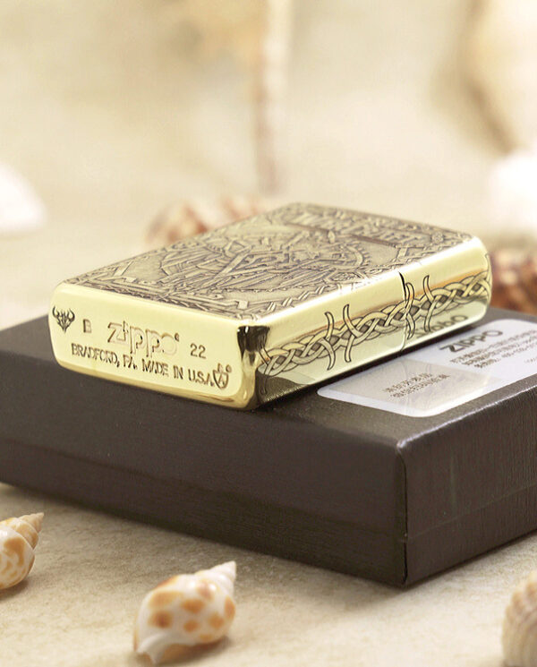 KZ217-05 Gold Zippo lighter with an ornate etched pattern rests on a black box, seaside shells scattered around.