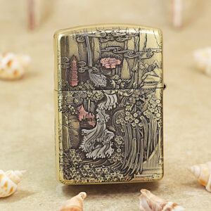 KZ218-01 Gold engraved lighter featuring a detailed Asian landscape with pagodas, trees, and flowing water, set on a sandy surface with seashells nearby.