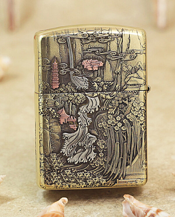 Gold engraved lighter featuring a detailed Asian landscape with pagodas, trees, and flowing water, set on a sandy surface with seashells nearby.
