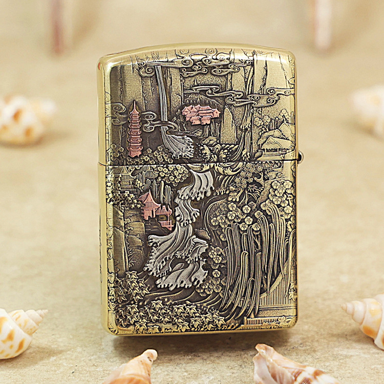 Gold engraved lighter featuring a detailed Asian landscape with pagodas, trees, and flowing water, set on a sandy surface with seashells nearby.