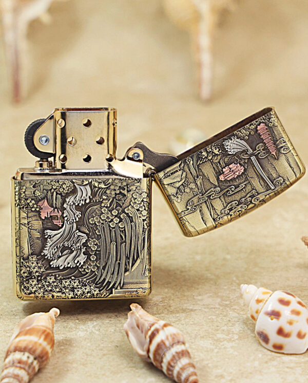 Engraved brass Zippo lighter, open lid, featuring intricate woodland scene with birds and trees on a sandy surface with seashells nearby﻿