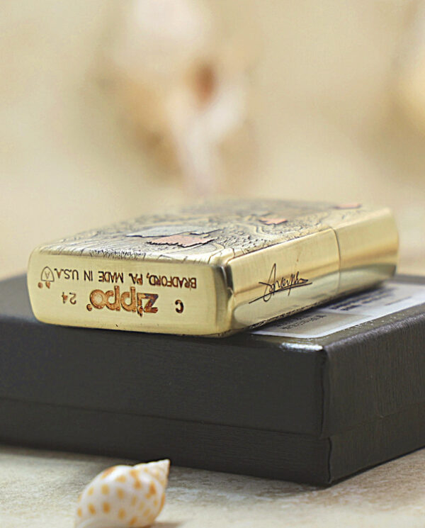 Gold Zippo lighter with engraved detailing placed on a black box, with blurred seashells in the soft background.