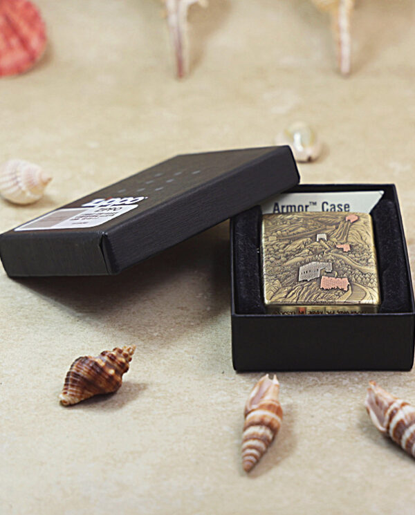 Gold-toned lighter in an open black box labeled Armor Case, resting on sand with seashells around it (product photo).