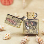 Gold Zippo lighter with a Tom and Jerry illustration reading 'LUCKIN' on the front, open to reveal the lid, placed on sandy surface among seashells.