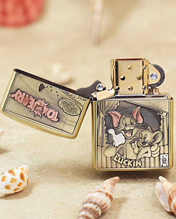 Gold Zippo lighter with a Tom and Jerry illustration reading 'LUCKIN' on the front, open to reveal the lid, placed on sandy surface among seashells.