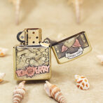 Tom and Jerry-themed brass Zippo lighter on a sandy beach, lid open, surrounded by small seashells on the surface.