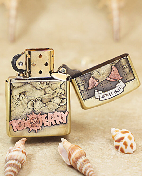 Tom and Jerry-themed brass Zippo lighter on a sandy beach, lid open, surrounded by small seashells on the surface.