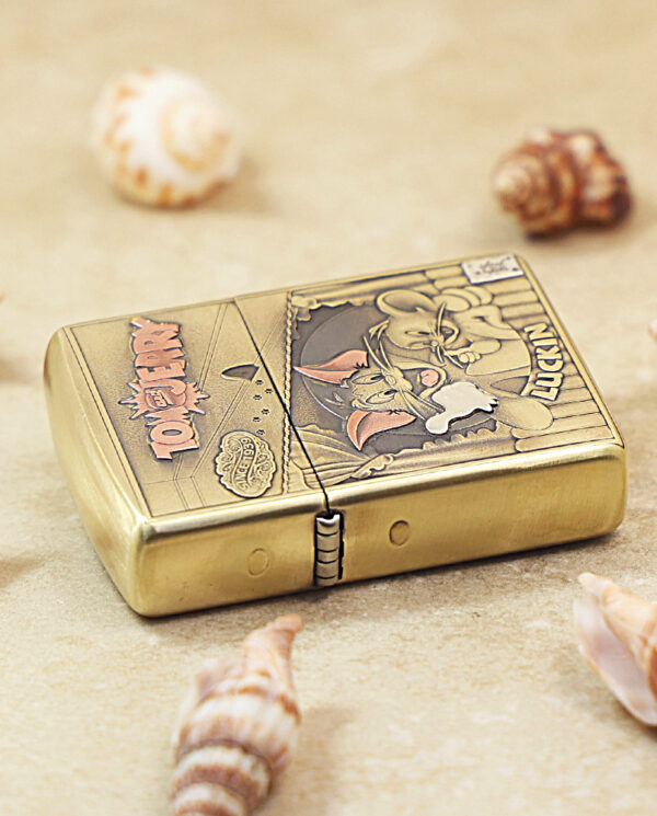 Gold Zippo lighter with a vintage cartoon graphic rests on sand among seashells on a beach.