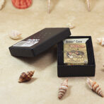 Gold-tone Zippo lighter with a Tom and Jerry scene inside a black Armor Case box, opened to reveal the design, on a sandy surface with seashells nearby.