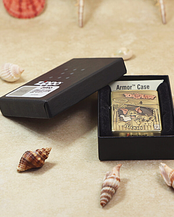 Gold-tone Zippo lighter with a Tom and Jerry scene inside a black Armor Case box, opened to reveal the design, on a sandy surface with seashells nearby.