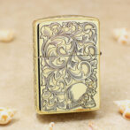 Gold metal lighter with ornate scrolling patterns and a skull at the bottom, placed on a sandy surface with seashells nearby.