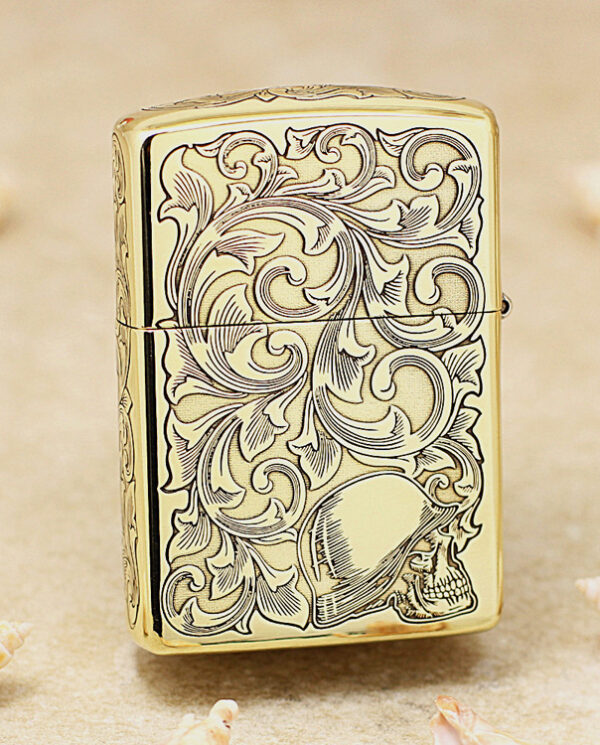 Gold metal lighter with ornate scrolling patterns and a skull at the bottom, placed on a sandy surface with seashells nearby.