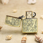 Gold-tone engraved Zippo-style lighter, open, resting on sand with seashells nearby.