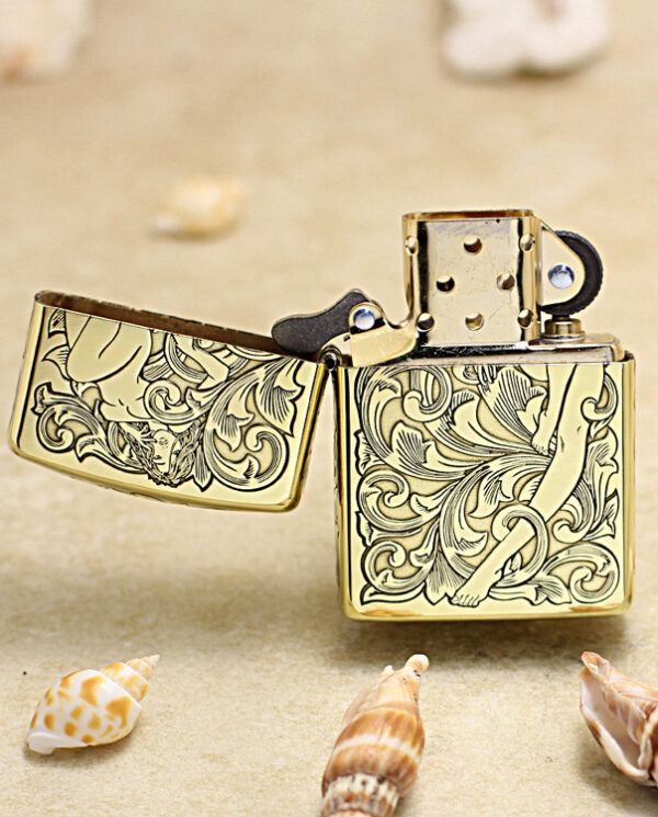 Gold-tone engraved Zippo-style lighter, open, resting on sand with seashells nearby.