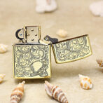 Gold engraved Zippo-style lighter with intricate scrollwork and a skull motif, placed on sand with seashells nearby.