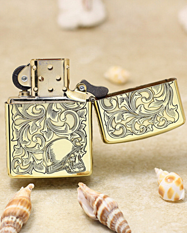Gold engraved Zippo-style lighter with intricate scrollwork and a skull motif, placed on sand with seashells nearby.