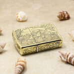 Gold-toned Zippo-style lighter with intricate floral engraving, resting on sand among seashells.