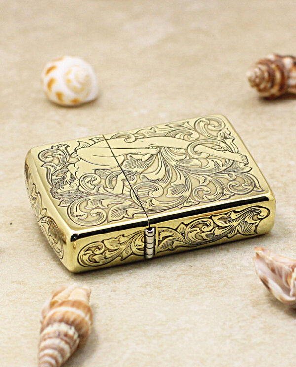 Gold-toned Zippo-style lighter with intricate floral engraving, resting on sand among seashells.