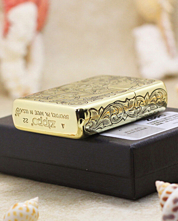 Gold engraved Zippo-style lighter resting on a black box, with seashells in the foreground.