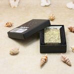 Black Zippo lighter with gold floral engraving in an open black box on a sandy surface with seashells nearby.